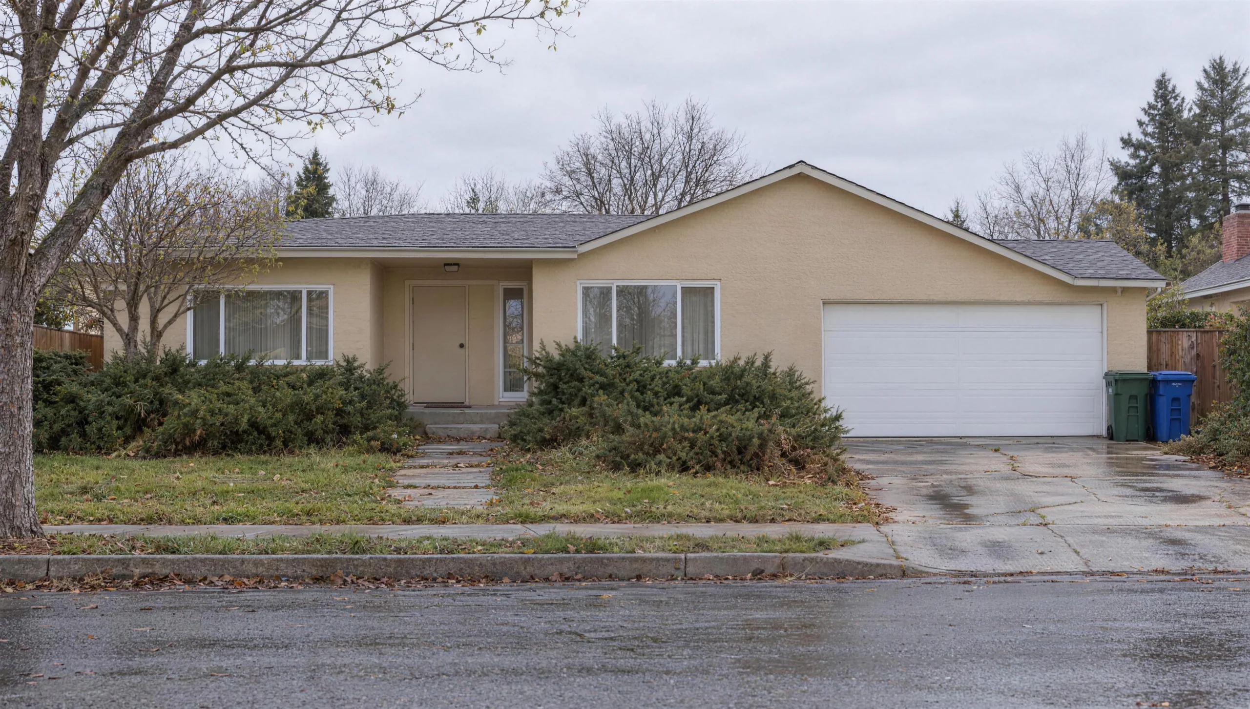 The same house before the exterior remodel on a rainy winter day: beige stucco, overgrown junipers and a cracked driveway